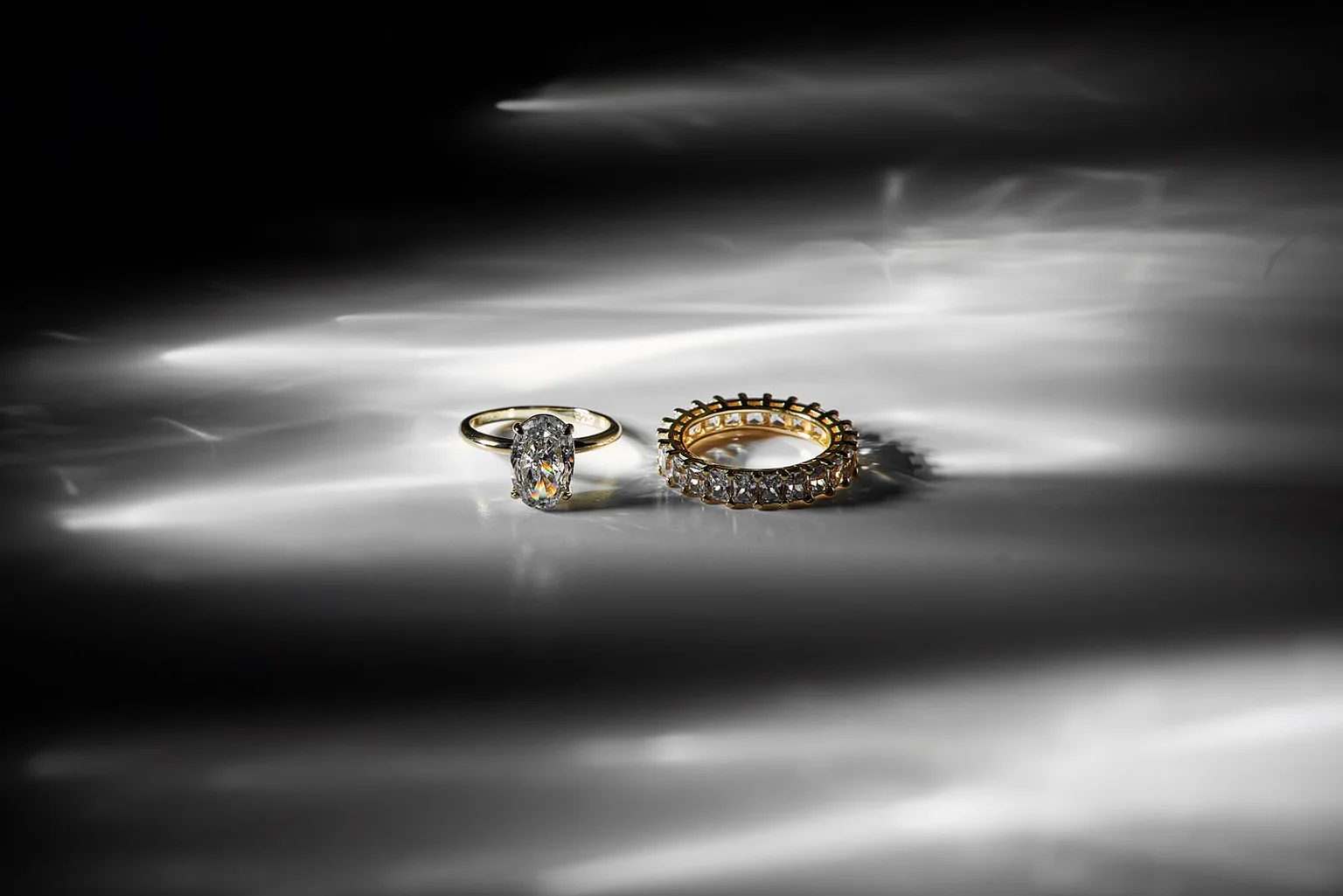 Two gold and diamond rings resting in natural light on a gray reflective surface